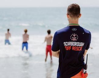 A lifeguard watches lifeguard qualification candidates standing in the surf while wearing a t-shirt that says "Scarborough Surf Rescue"