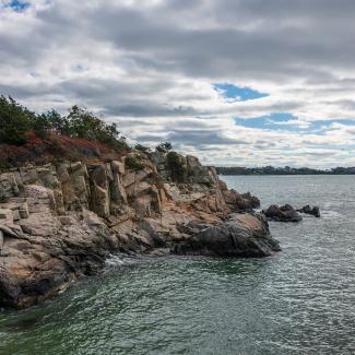 The rocky shoreline of Fort Wetherill State Park meets the calm waters of Narragansett Bay as a blue sky peeks out behind the clouds on a cold day and tranquil day