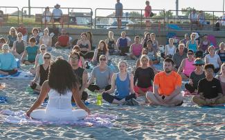 A large group of people sit in a yoga pose on a sandy beach