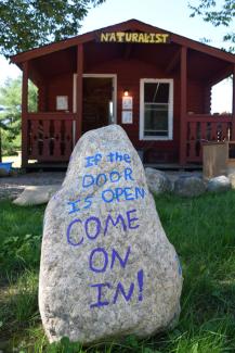 A painted rock reads, "Come on in!" in front of a naturalist cabin