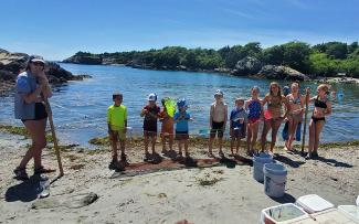 A school group of elementary-aged kids stands along the shoreline with a siene net