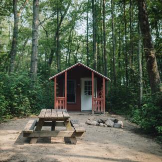 A rustic cabin with a small window and porch near a circular rock campfire and picnic table among the tall trees and green brush at Burlingame State Campground