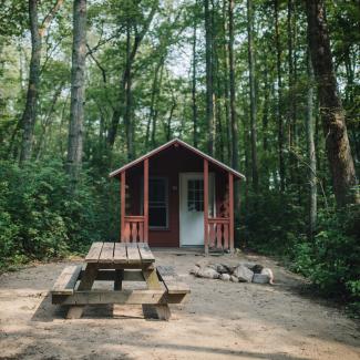 A rustic cabin with a small window and porch near a circular rock campfire and picnic table among the tall trees and green brush at Burlingame State Campground