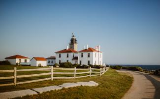 The white with red roofed historic Beavertail lighthouse sits on a grassy green lawn with bright blue sky