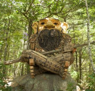 Art installation of a troll built out of reclaimed wood including whiskey barrels sitting on a boulder in the woods